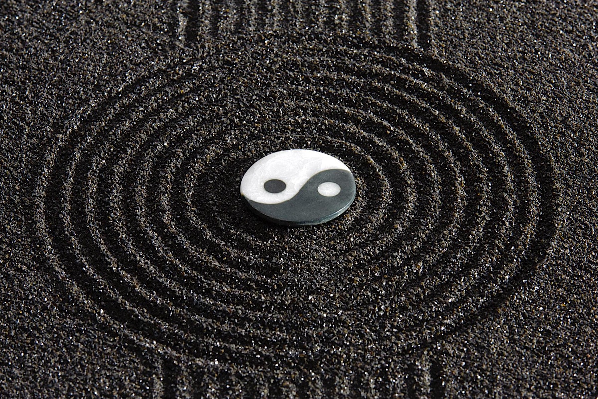 Ein Yin-Yang-Symbol, halb weiß und halb dunkel, liegt im Zentrum einer Reihe konzentrischer Kreise, die in dunklen Sand geätzt sind. Ähnlich wie beim Yin-Yoga bilden die Kreise ein harmonisches Muster um das Symbol und vermitteln ein Gefühl von Ausgeglichenheit und Ruhe.
