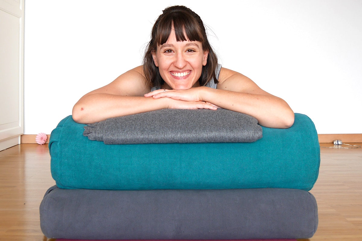 Katharina Balande lächelt und legt ihr Kinn auf einen Stapel aus drei gefalteten Yoga-Bolstern in Blaugrün, Grau und Dunkelgrau auf einem Holzboden vor einer schlichten weißen Wand. Sie wirkt zufrieden.