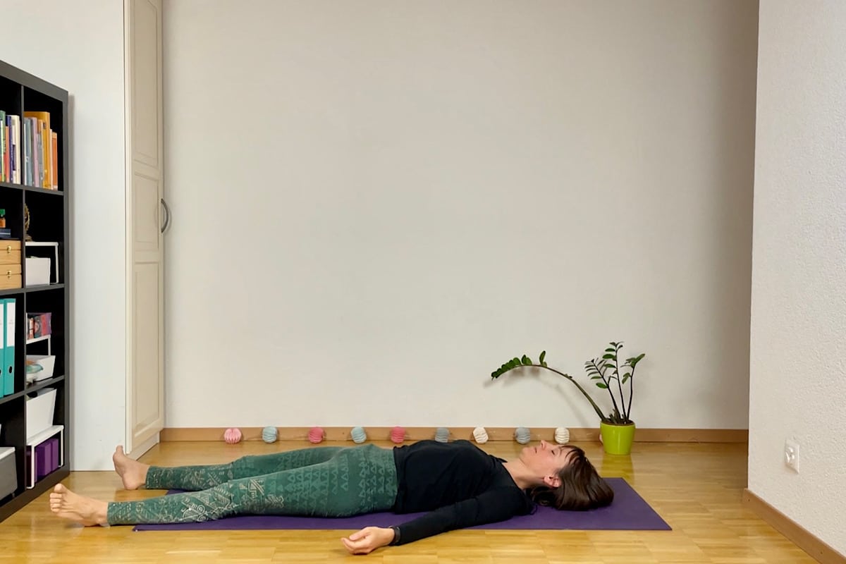 Eine Person liegt auf dem Rücken auf einer Yogamatte und übt Shavasana. Der hell erleuchtete Raum eignet sich mit seinem Holzboden, dem Bücherregal links und einer kleinen Pflanze mit Dekoartikeln, die an der Wand für Ruhe sorgen, perfekt für eine Yin Yoga Sequenz für den Herbst.