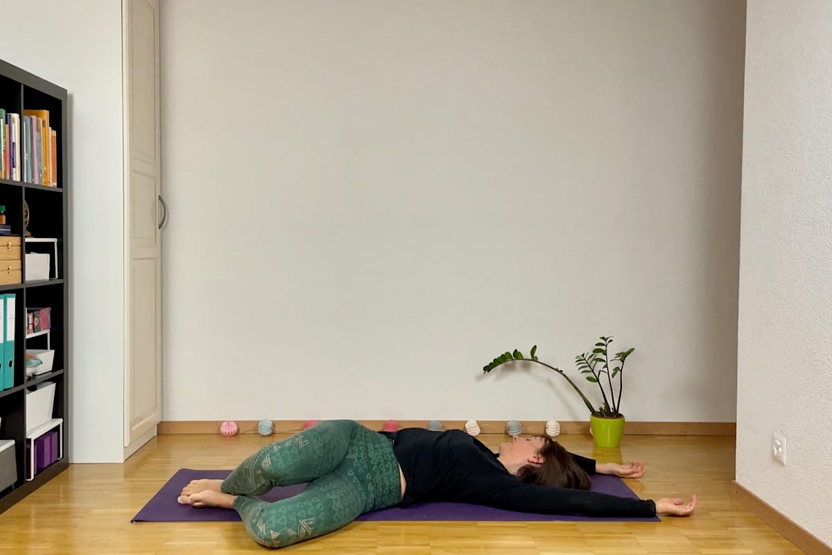 Eine Person liegt auf einer violetten Yogamatte und ist in eine beruhigende Yin-Yoga-Sequenz für den Herbst vertieft. In dem geräumigen Raum führt sie eine sanfte Drehung aus, die Knie sind gebeugt und die Beine auf einer Seite. Die Arme sind neben der kleinen Pflanze und dem Bücherregal ausgestreckt, die diese friedliche Umgebung vervollständigen.