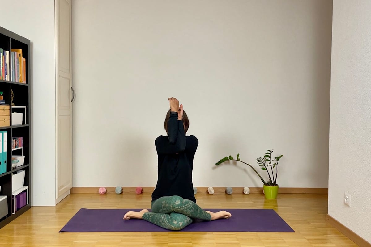 Eine Person praktiziert die Yin Yoga Stellung "Adlerarme" auf einer violetten Matte in einem minimalistischen Raum und übt einen Teil der Yin Yoga Sequenz für den Herbst. Sie sitzt in einer Taubenhaltung mit nach oben ausgestreckten Armen, im Hintergrund sind Regale mit Büchern und eine kleine Pflanze zu sehen.