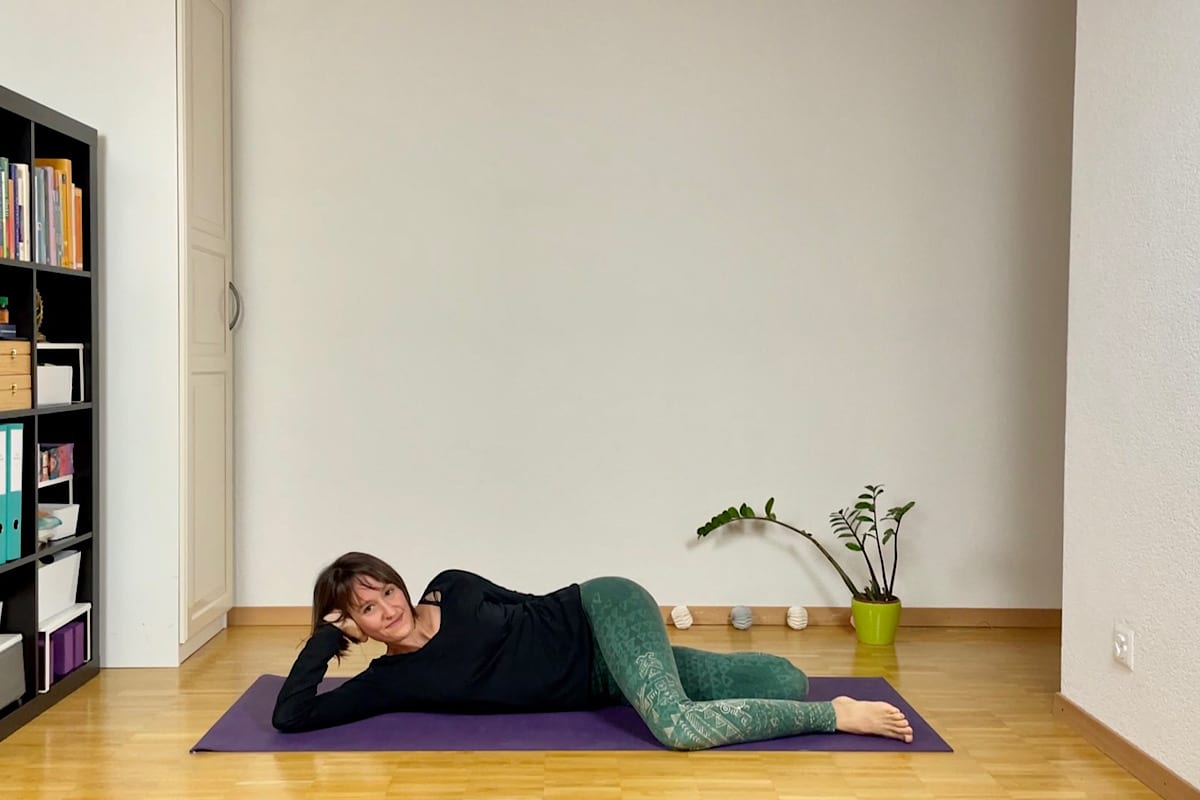 Eine Person liegt seitlich auf einer Yogamatte und führt eine Yin-Yoga-Sequenz für den Herbst aus. Hier: die liegende Katze. In dem hellen Raum mit Holzboden stützt eine Hand den Kopf, während die andere auf der Matte ruht. Ein Bücherregal und eine Zimmerpflanze verleihen der ruhigen Umgebung eine einladende Note.