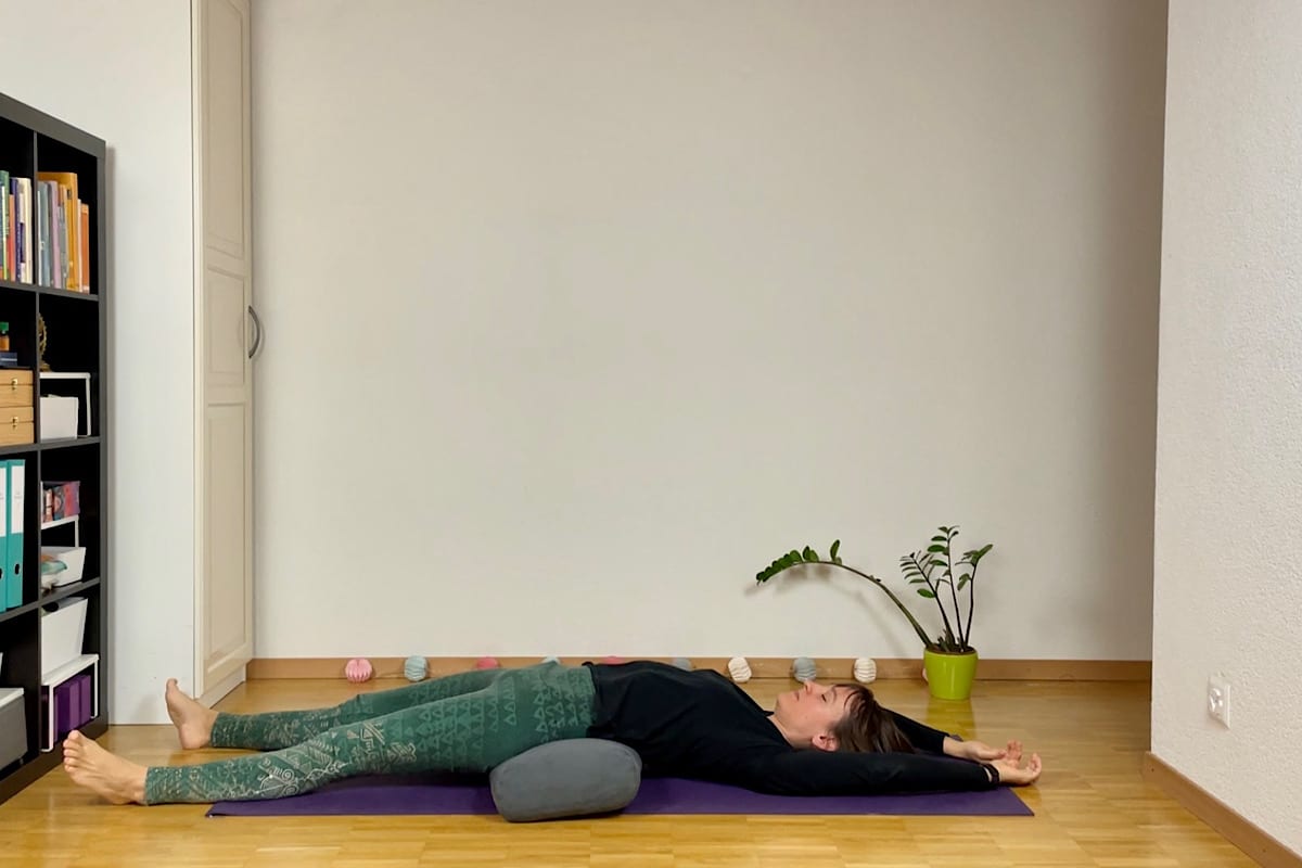 Eine Person liegt auf dem Rücken auf einer Yogamatte und führt eine beruhigende Yin-Yoga-Sequenz für den Herbst aus. Ihre Arme sind über dem Kopf ausgestreckt, mit einem Kissen unter den Knien - die Regenbogenbrücke. Sie trägt ein schwarzes Oberteil und gemusterte grüne Hosen und entspannt sich neben einer kleinen Pflanze und einem mit Büchern gefüllten Regal auf der linken Seite.