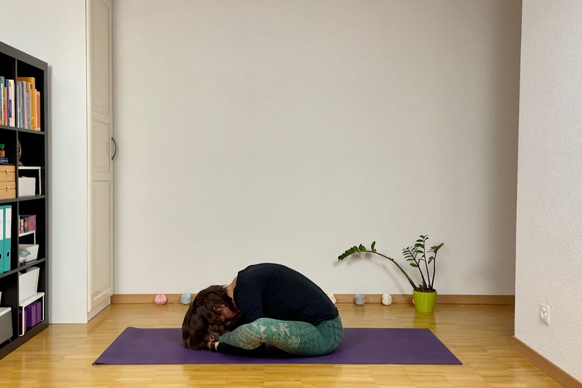 Eine Person praktiziert Yin Yoga in einer sitzenden Vorwärtsbeuge, der Schildkröte, auf einer lila Matte mit gekreuzten Beinen. Der Holzboden des Raums ergänzt die beruhigende Atmosphäre dieser Herbstsequenz mit einem Bücherregal auf der linken Seite, einer kleinen Pflanze auf der rechten Seite und dekorativen Gegenständen vor einer schlichten weißen Wand.