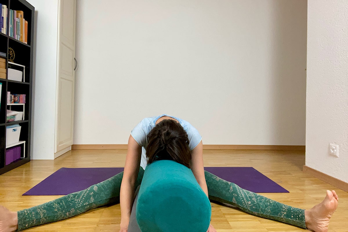 Eine Person praktiziert anmutig Yin Yoga in einem ruhigen Raum mit hellem Holzboden und lässt sich vom belebenden Geist des Frühlings anstecken. Sie sitzt mit gespreizten Beinen und beugt sich nach vorne über ein grünes Polster auf einer violetten Matte. Ein Bücherregal auf der linken Seite trägt zur ruhigen Atmosphäre bei.