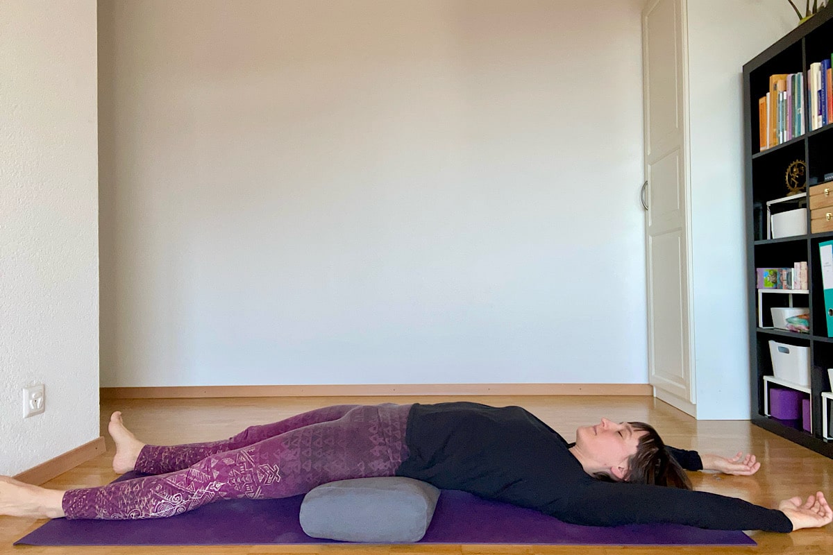 Katharina Balande in der Yin Yoga Stellung Regenbogenbrücke. Rückenlage, Beine leicht geöffnet, Arme über den Kopf, Bolster unter dem Becken.