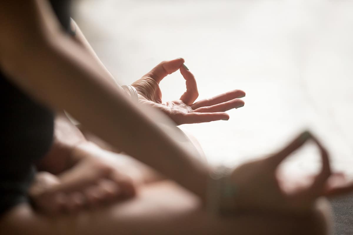 Nahaufnahme von zwei Personen, die im Schneidersitz in einer Meditationshaltung sitzen und Wechselatmung praktizieren. Die Hände ruhen auf den Knien und die Finger befinden sich in einer Mudra-Position. Der sanft unscharfe Hintergrund verstärkt die friedliche und konzentrierte Atmosphäre der Szene.