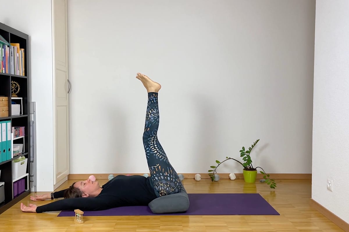 Eine Person liegt in einem Raum auf dem Rücken auf einer lila Yogamatte, die Beine sind gerade nach oben gestreckt und die Arme sind seitlich ausgestreckt.