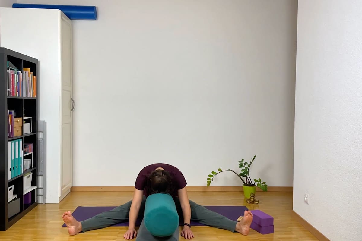 Eine Person übt in einer Yin-Yoga-Sitzung eine Vorwärtsbeuge mit gespreizten Beinen, wobei ein Yoga-Bolster ihren Oberkörper stützt.