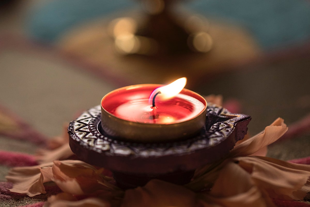 Eine kleine rote Wachskerze brennt in einem aufwendig gemusterten Halter, umgeben von weichen Blütenblättern. Als ob sie eine magische Auszeit spenden würde, wirft die Flamme ein warmes Licht, mit verschwommenen Farben im Hintergrund, die die gemütliche Atmosphäre verstärken.