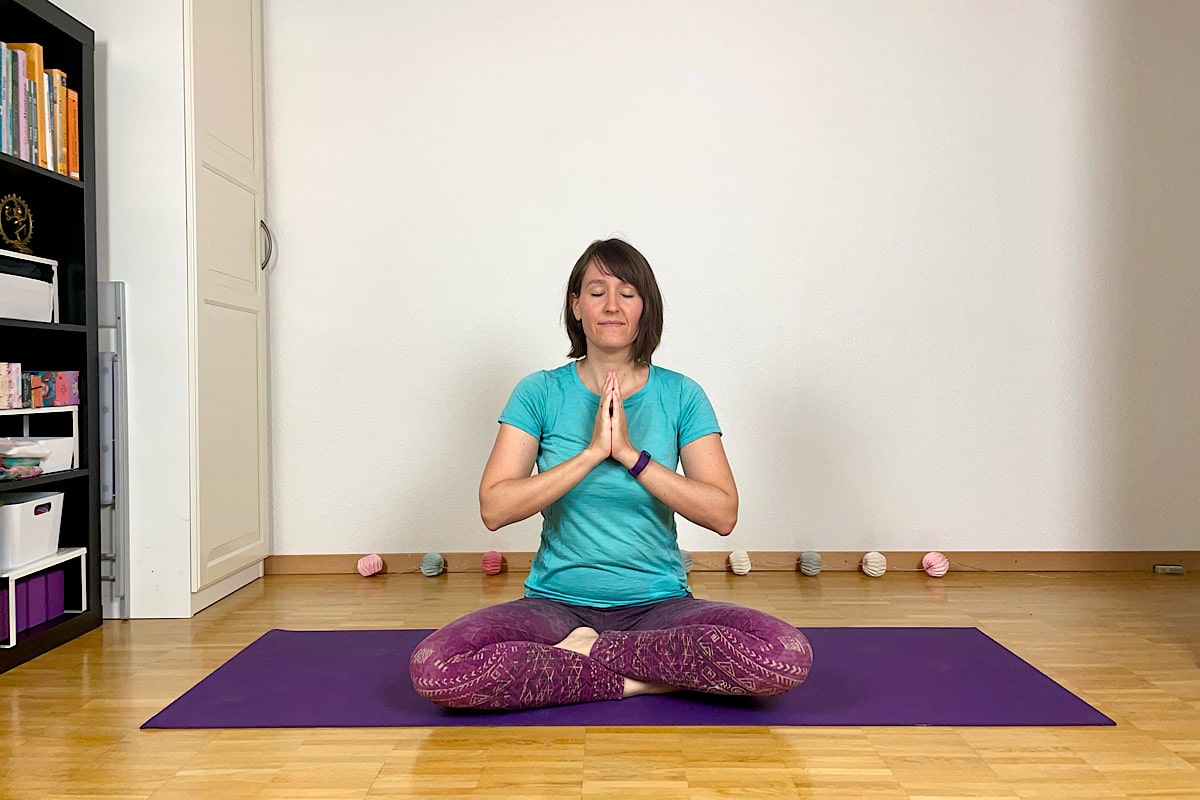 Eine Person sitzt im Schneidersitz auf einer lila Yogamatte und erlebt eine magische Auszeit, während sie mit den Händen in Gebetshaltung meditiert. Sie trägt ein blaugrünes Hemd und lila Leggings. Im Hintergrund ist ein Regal mit Büchern und Wollknäueln zu sehen, die auf dem Boden verstreut sind und Ruhe zwischen den Welten bieten.
