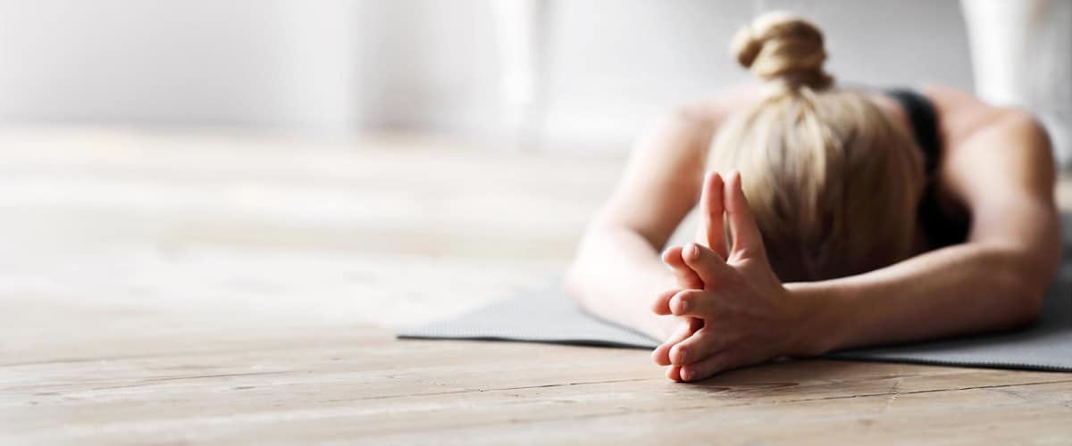 Eine Person mit blondem Haar, das zu einem Knoten gebunden ist, streckt sich auf einer Yogamatte und genießt eine magische Auszeit. In der Stellung des Kindes strecken sich ihre Arme nach vorne über den Holzboden. Der Fokus bleibt auf ihren Händen vor dem unscharfen Hintergrund, was ein Gefühl von Frieden und Ruhe vermittelt.