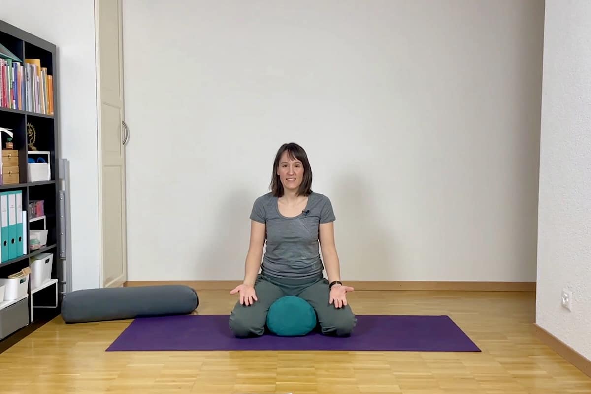 Eine Frau sitzt im Schneidersitz auf einer Yogamatte. Unter ihr liegt ein blaugrünes Kissen, ihre Hände ruhen auf den Knien. Neben ihr sind in einem aufgeräumten, lichtdurchfluteten Raum ein Bücherregal und ein Nackenkissen zu sehen.