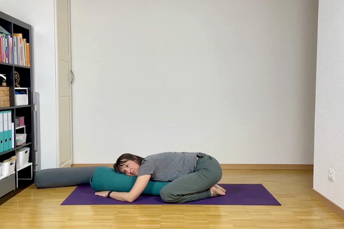 Eine Person liegt in der entspannten Stellung eines Kindes auf einer Yogamatte und hält ein Nackenkissen unter Brust und Kopf. In der Nähe befindet sich in einem aufgeräumten Raum mit Bücherregalen ein weiteres Nackenkissen.