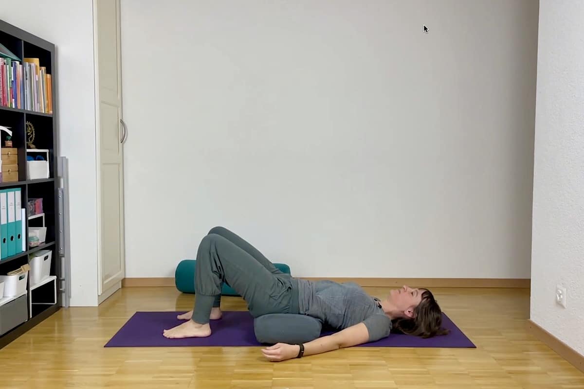 Eine Frau liegt mit angewinkelten Knien und den Füßen auf dem Boden auf einer Yogamatte. Unter ihren Hüften befindet sich ein Kissen. Ihre Arme ruhen seitlich an den Seiten, die Handflächen zeigen nach oben. In einem hellen, aufgeräumten Raum mit Regalen und Büchern.