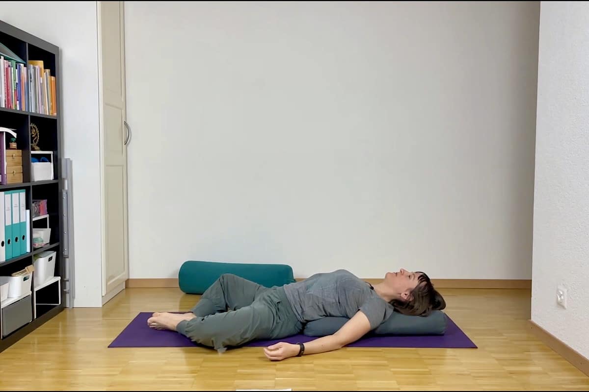 Eine Person liegt mit nach außen gebeugten Knien auf dem Rücken auf einer Yogamatte, gestützt durch Kissen unter Oberkörper und Knien, in einem ruhigen Raum mit Bücherregalen und Holzboden.