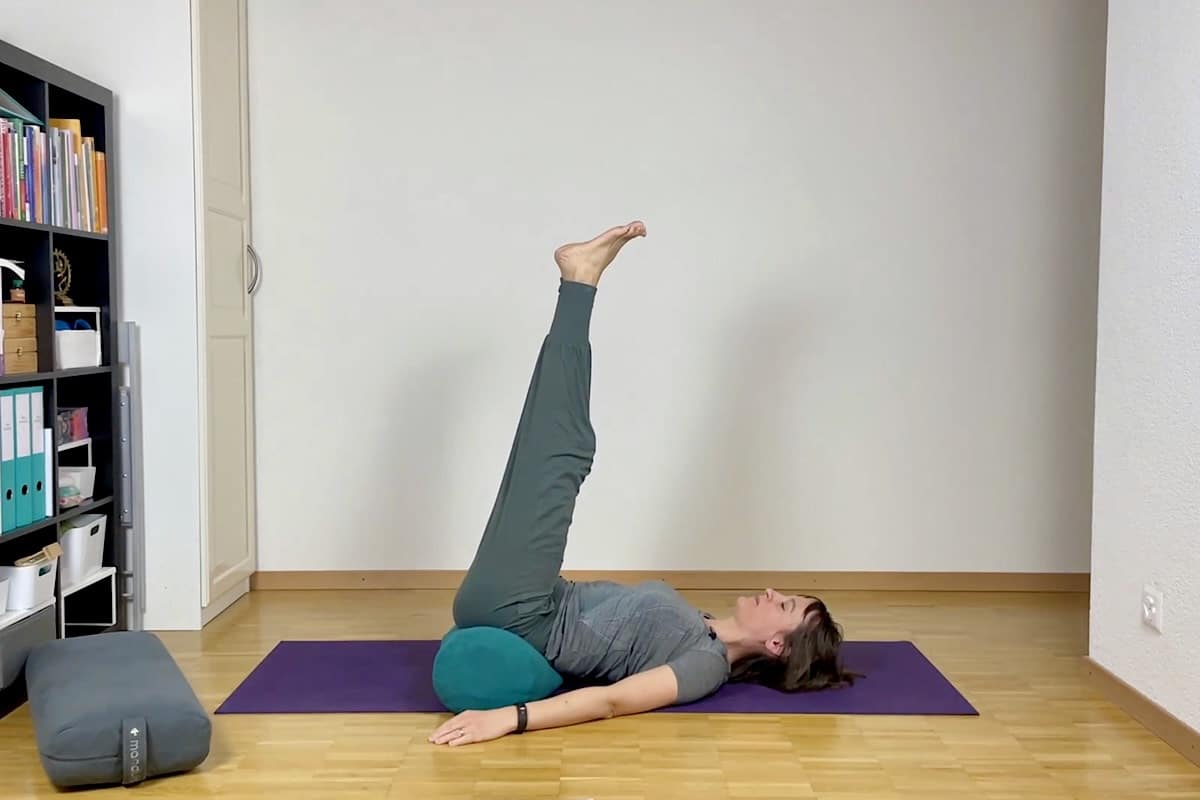 Eine Frau liegt mit ausgestreckten Beinen auf dem Rücken auf einer Yogamatte, gestützt durch ein Kissen unter ihrem unteren Rücken, in einem minimalistischen Raum mit Regalen und Büchern.
